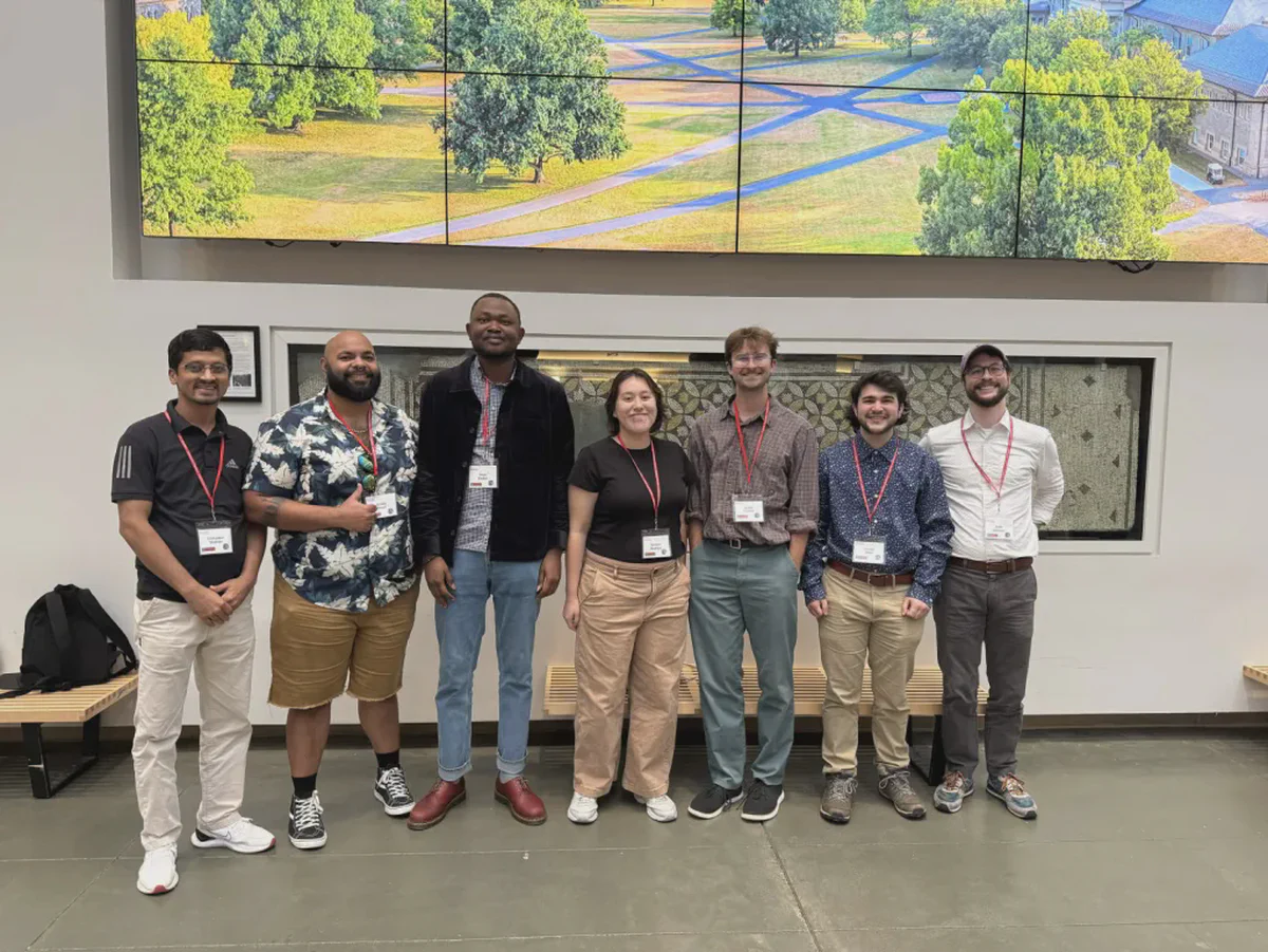 Seven conference attendees wearing name badges pose together beneath a large campus display.